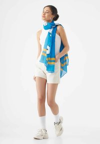 Lichtblauwe sjaal met speelse patronen, casual gedrapeerd, aangevuld met crèmekleurige shorts en witte sokken met sportieve schoenen.