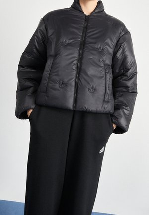 Osoba w czarnej puchowej kurtce z zamkiem błyskawicznym i czarnych spodniach dresowych Adidas, z rękami w kieszeniach, stojąca na jednolitym tle.