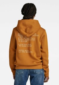 Sudadera color mostaza con capucha ajustable. Presenta un gráfico de texto blanco en la parte posterior, fabricada con una tela suave y un corte relajado.