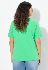 T-shirt verde a maniche corte con una vestibilità rilassata e maniche raglan, realizzata in un tessuto morbido. Indossato con jeans blu in denim, visto da dietro.