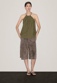 Olivově zelený halter top s sklady, doplněný hnědými semišovými kraťasy do kolen. Modelka stojí s rukama v kapsách, má na sobě ploché černé sandály.