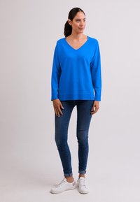 Blauer V-Ausschnitt-Pullover mit langen Ärmeln, lässiger Schnitt, über enganliegenden Denim-Jeans und weißen Sneakers getragen, vor einem einfarbigen hellen Hintergrund.