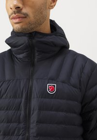 Sötétkék párnázott kabát magas gallérral és cipzáras záródással. Kiemelkedő logó emblémával és vízszintes steppelési mintával rendelkezik.