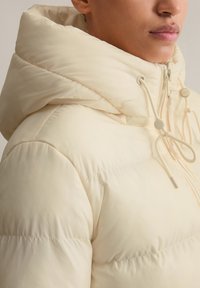 Aufblasbare Jacke in hellem Creme mit Kapuze. Verfügt über einen Reißverschluss, Kordelzüge und eine gesteppte Textur mit sichtbaren Nähten.