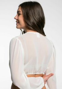Witte doorzichtige blouse met lange pofmouwen, gathered taille en textuurstof. Achteraanzicht toont soepele contouren en subtiele drapering.