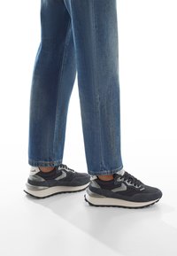 Gambe che indossano jeans blu e sneakers nere e grigie, in piedi su una superficie bianca con riflessi di luce.