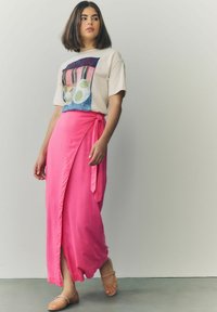 Vit t-shirt med en färgglad grafik på framsidan, kombinerad med en ljusrosa omslagskjol med knytdetalj och sidslits. Platta sandaler.