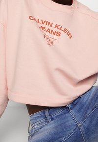Sudadera corta rosa con "CALVIN KLEIN JEANS 1978" impreso en naranja oscuro. Presenta un escote redondeado y una tela suave. Combinada con jeans azules.