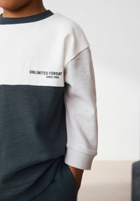 Felpa color block con design bianco e grigio scuro, caratterizzata dal testo "UNLIMITED FUNDAY" e "SINCE 1982" in nero. Materiale misto cotone.