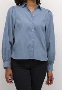 Personne portant un chemisier bleu clair à manches longues bouffantes et un pantalon noir, debout devant un fond clair uni.