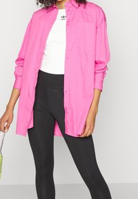 Chemise rose vif à manches longues, dotée d'une poche poitrine, portée par-dessus un haut blanc et un legging noir. Détails en chaîne sur le sac à main.