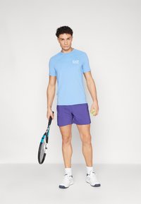 Heledad helesinine spordipolt, millel on valge logo, tumepunaste lühikesed püksid. Käes on tennisereket ja erks roheline tennisepall.