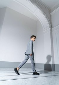 Szary garnitur o teksturowym wykończeniu, czarne buty i rozczochrane włosy; chłopak w ruchu na tle minimalistycznym, jasnym.