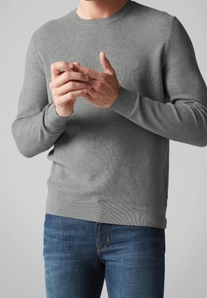 Maglione grigio lavorato a maglia con scollo rotondo, polsini e orlo a costine. Indossato con jeans di denim blu, evidenziando i dettagli del tessuto testurizzato.
