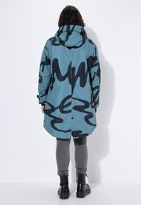 Cappotto impermeabile blu con motivo astratto nero, cappuccio, maniche lunghe e orlo curvo, indossato sopra pantaloni grigi e stivali neri.