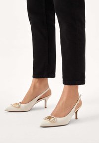 Beżowe skórzane buty na obcasie typu slingback z ostrym noskiem, ozdobione złotą klamrą z akcentami kryształów i smukłą konstrukcją obcasa.