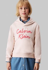 Felpa rosa chiaro con tasca frontale, decorata con testo rosso in rilievo "Calvin Klein". Materiale morbido, vestibilità comoda e polsini a coste. Gonna di jeans sotto.