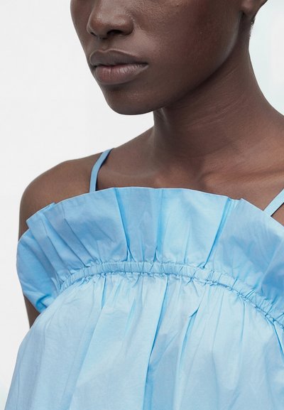 Κοντινό πλάνο ενός ατόμου που φοράει ένα ανοιχτό μπλε τοπ με βολάν και λεπτές τιράντες, δείχνοντας τον λαιμό, τους ώμους και τα χείλη του.