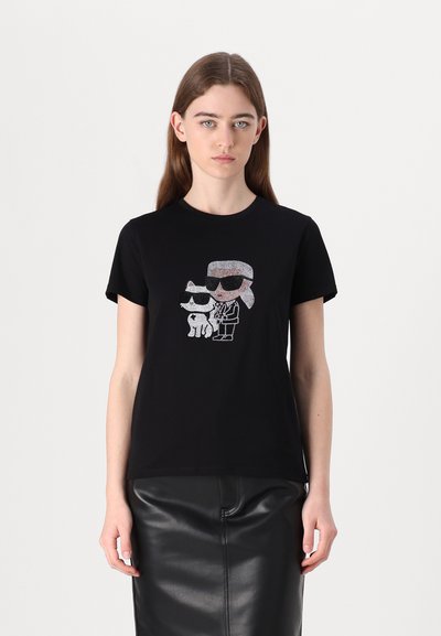 Jeune femme portant un t-shirt noir avec une figure de cartoon en strass représentant une personne et un chien portant des lunettes de soleil, associé à une jupe en cuir noire.