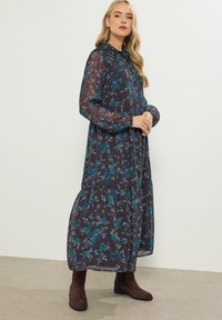 Abito maxi floreale in tessuto scuro con sfumature di blu, dotato di colletto, maniche lunghe e design a strati. Abbinato a stivali alla caviglia marroni.