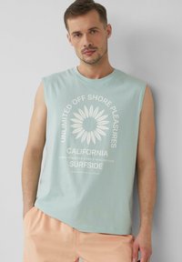 Hellblaues, ärmelloses T-Shirt mit weißem Grafiktext und einem Blumendesign, aus Baumwolle, kombiniert mit pfirsichfarbenen Shorts.