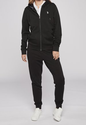 Persona vestida con sudadera negra con cremallera, pantalones jogger negros, camiseta blanca debajo y zapatillas blancas, de pie frente a un fondo claro y liso.