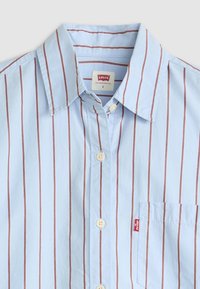 Chemise bleu clair à boutons avec des rayures verticales rouges et blanches, col classique, poche sur la poitrine gauche, et étiquette logo sur la poche.