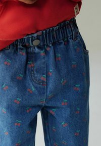 Pantalones de mezclilla azul con un estampado de cerezas en rojo y verde, cintura elástica con botón metálico y costuras en contraste.