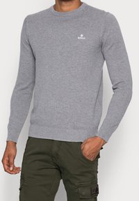 Pull gris en maille avec un col rond, des poignets et un ourlet côtelés, et un petit logo blanc sur la poitrine. Porté avec un pantalon vert olive.