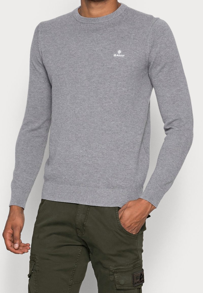 Pull gris en maille avec un col rond, des poignets et un ourlet côtelés, et un petit logo blanc sur la poitrine. Porté avec un pantalon vert olive.