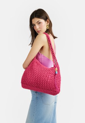 Giovane donna con top rosa e jeans che porta una grande borsa a tracolla rosa intrecciata con perline decorative sulla tracolla.