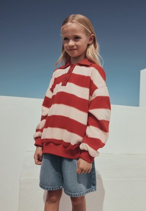 Ung pige står udendørs iført en rød- og hvidstribet sweater og cowboyshorts mod en klar blå himmel.