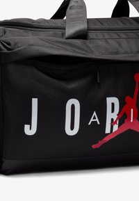 Sort duffel taske med en tekstureret overflade, prydet med store hvide bogstaver der staver "J O A" og en rød silhuet af en springende figur.