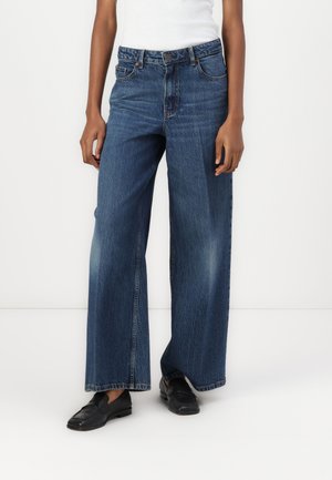 Persoon draagt een broek met hoge taille, brede pijpen van blauwe spijkerstof met lichte vervaging en zwarte loafers, staand tegen een effen witte achtergrond.