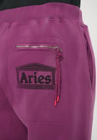 Ljubičaste trenirke s prednjim džepom na zatvarač, crnim "Aries" logotipom i kontrastnim crvenim šnuralama. Izrađene su od mekog, teksturiranog materijala.