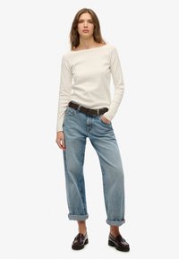 Off-White Langarmshirt kombiniert mit hellblauen, leicht übergroßen Jeans. Die Jeans haben umgeschlagene Säume und sind mit einem braunen Gürtel befestigt. Braune Schuhe.