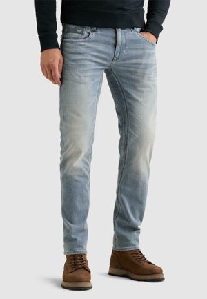 Homme portant un jean bleu clair délavé, des bottes marron à lacets et un haut noir à manches longues avec une main dans la poche.