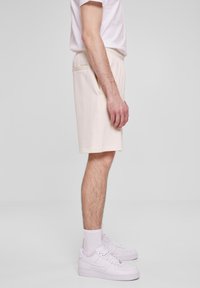 Homme portant un short beige clair, des chaussettes blanches et des baskets blanches, se tenant sur un fond gris uni, vu de côté.