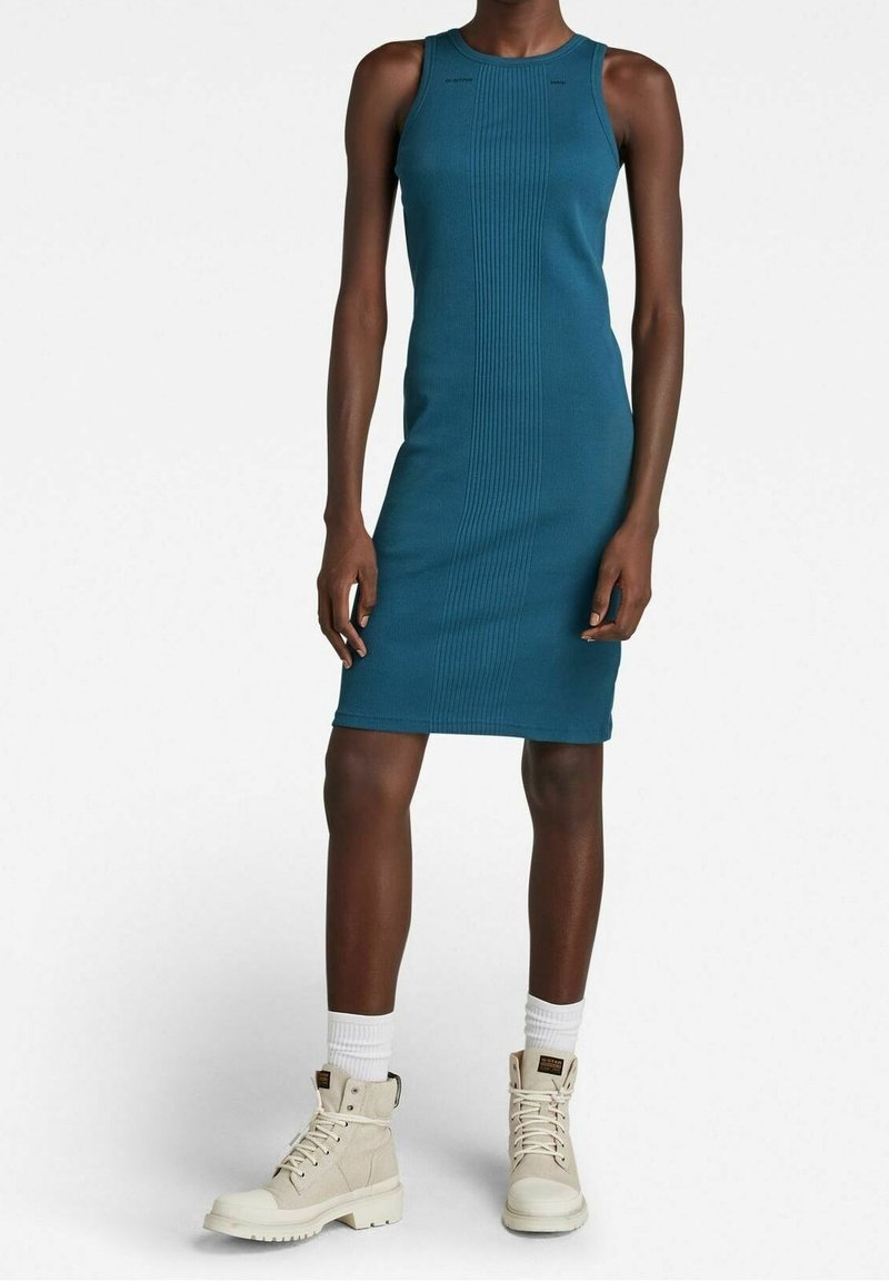 Tiefgrünes, geripptes Strickkleid in enganliegender Form, runder Ausschnitt und ärmellos, kombiniert mit beigen Knöchelstiefeln und weißen Socken.
