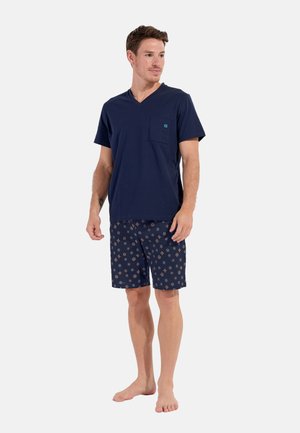 T-shirt en coton marine avec col en V et poche, associé à un short marine à motifs comprenant de petits dessins dorés et blancs. Position pieds nus.