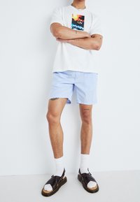 T-shirt blanche avec impression graphique, shorts rayés bleu clair, chaussettes blanches et sandales brunes avec une semelle distinctive. Design minimaliste.
