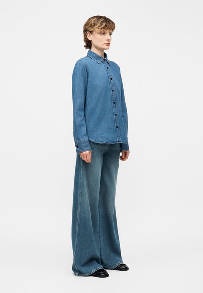 Άτομο που φοράει εφαρμοστό πουκάμισο από τζιν με κουμπιά και τζιν παντελόνι με φαρδιές σκάλες, στέκεται πλαγίως με μαύρα παπούτσια μπροστά σε απλό φόντο.