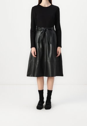 Kobieta ubrana w czarną bluzkę z długim rękawem, czarną skórzaną spódnicę do kolan z paskiem do wiązania, czarne skarpetki i czarne płaskie buty na białym tle.