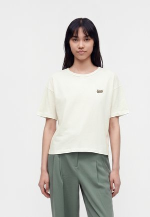 Tânără cu păr negru drept, purtând un tricou crem cu mâneci scurte cu textul "Sweet" și pantaloni verzi largi, stând în fața unui fundal alb.