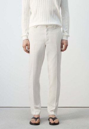 Osoba ubrana w kremowe, dopasowane spodnie, biały prążkowany sweter i czarne japonki, stojąca na tle jednolitego jasnego tła.