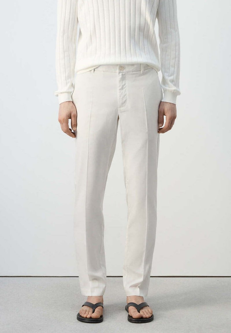 Personne portant un pantalon sur mesure écru, un pull blanc côtelé et des sandales tongs noires, debout devant un fond clair uni.