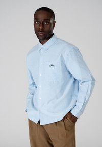 Homme portant une chemise à rayures bleu clair et blanc boutonnée avec un détail brodé sur la poche, et un pantalon marron, posant les mains dans les poches.