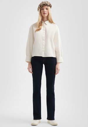 Femme blonde portant une chemise blanche boutonnée, un jean droit foncé, des chaussures blanches et un béret à motif léopard, debout devant un fond uni.