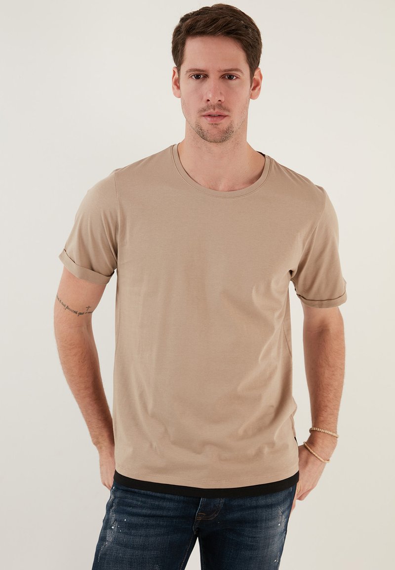 Beige Kurzarm-T-Shirt aus weicher Baumwolle, mit Rundhalsausschnitt und aufgerollten Ärmeln. Modell trägt Slim-Fit-Jeans und steht vor einem hellen Hintergrund.