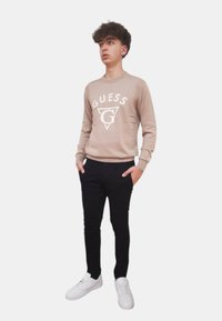 Maglione a maglia beige con logo "GUESS" sul petto, abbinato a pantaloni neri slim-fit e sneakers bianche. Il modello è in piedi con le mani in tasca.